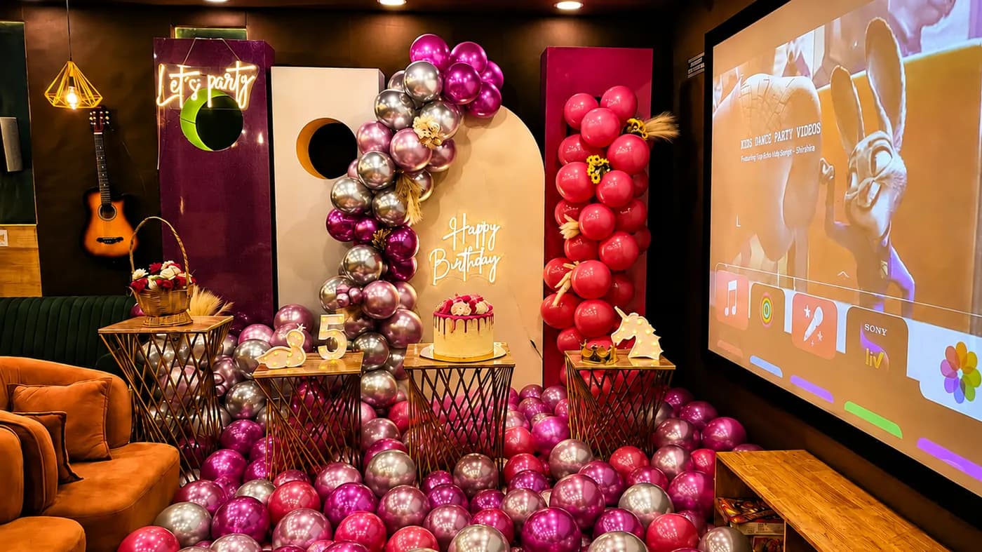 A pink, magenta and silver balloon arch and floor balloons around a cream backdrop with a neon Happy Birthday sign, a lit 5 sign and a drip cake, beside the projection screen and a guitar on the wall