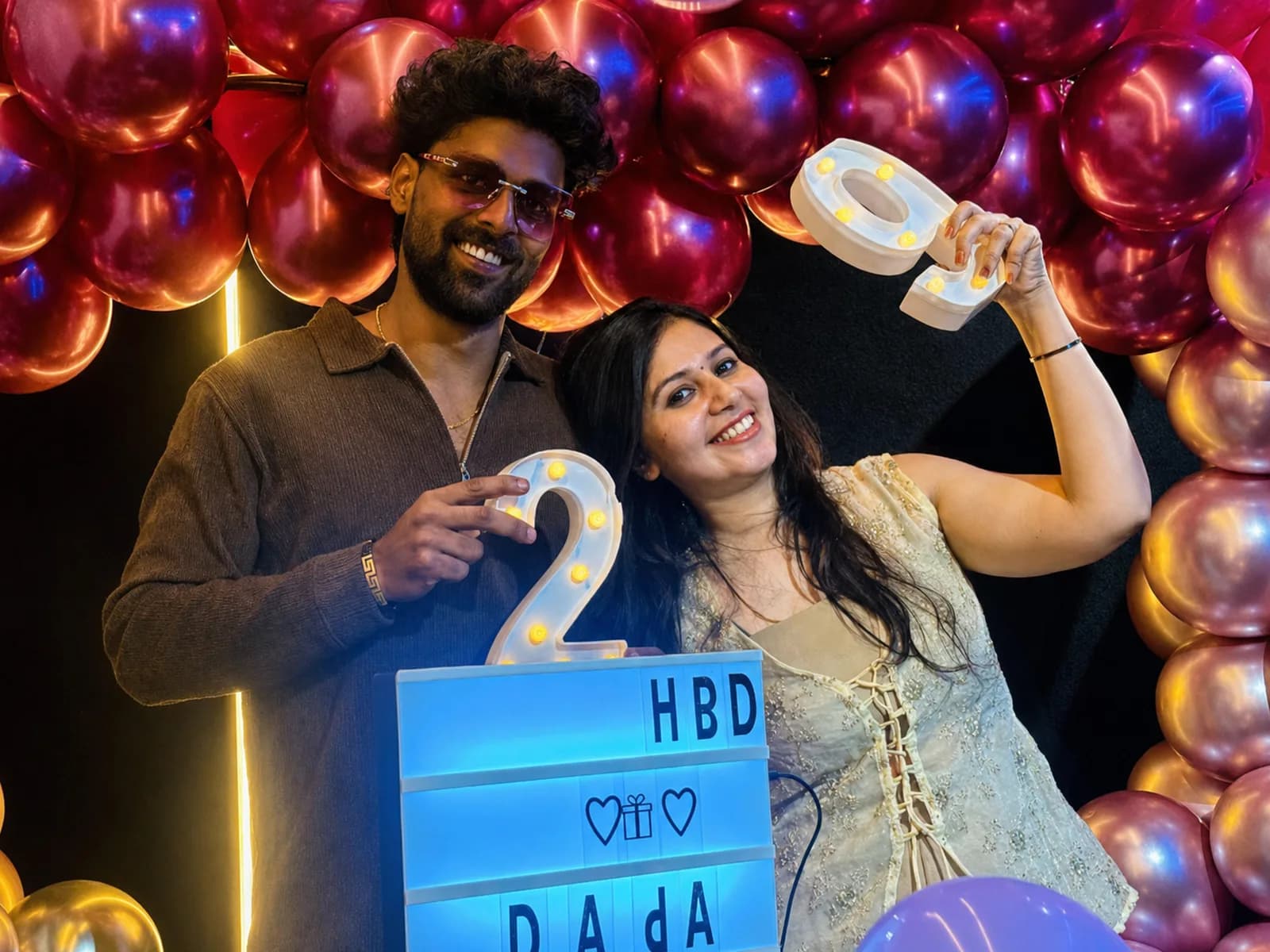 A couple with a lit HBD letter board under a red and gold chrome balloon arch
