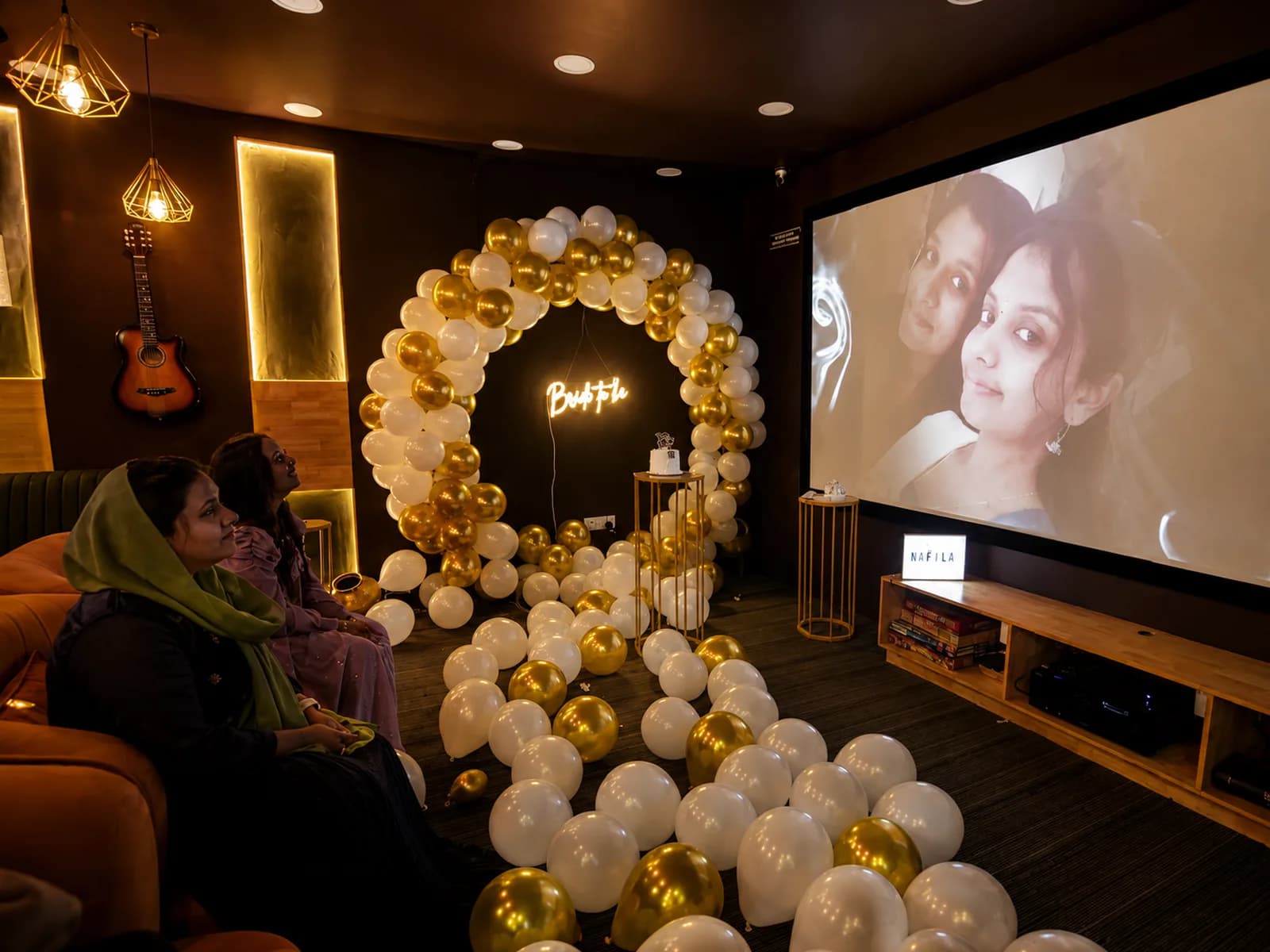 Guests on a sofa watching a film on the projection screen, beside a white and gold balloon ring with a neon Bride To Be sign