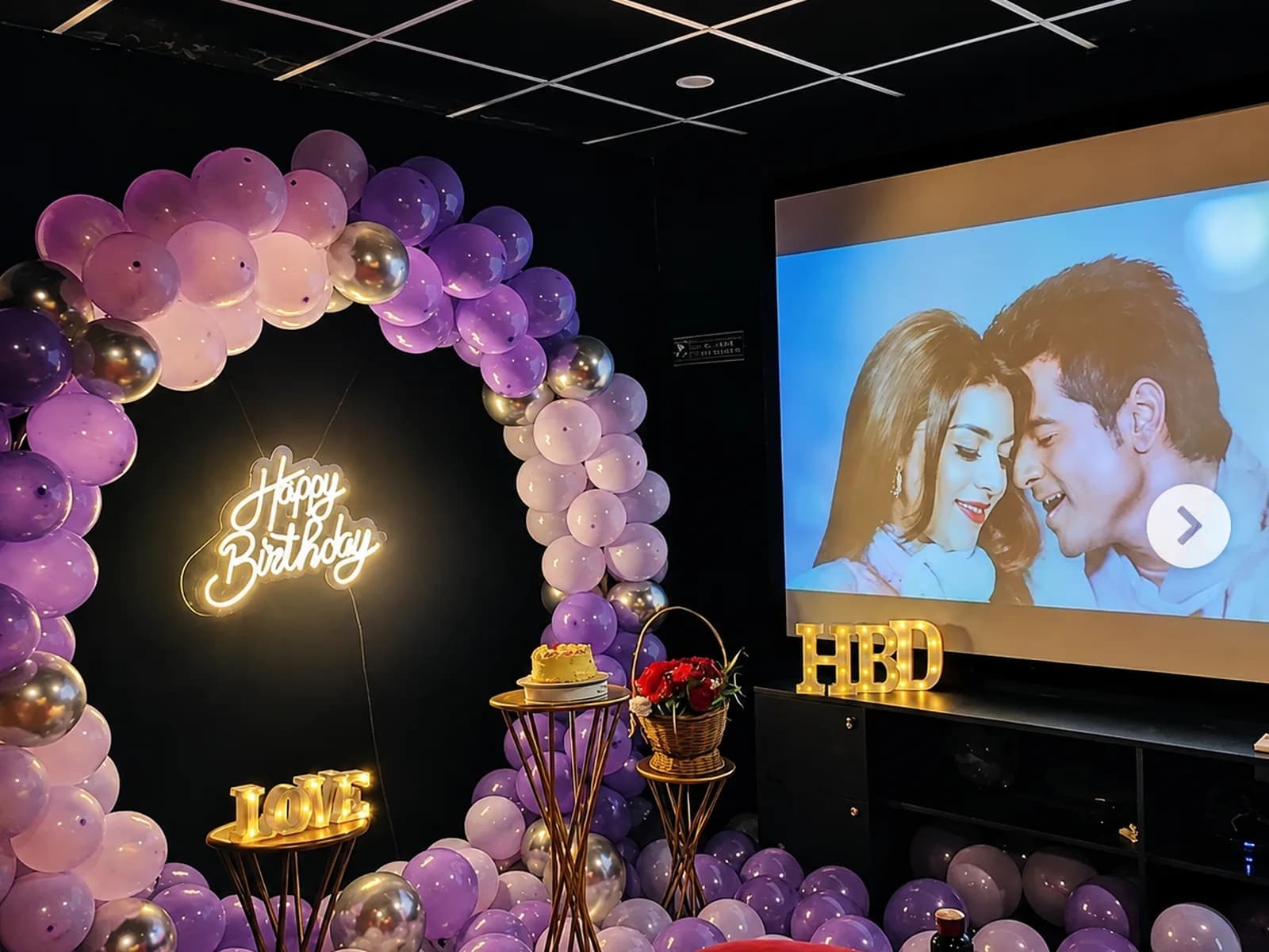 A purple and chrome balloon ring around a neon Happy Birthday sign, with a cake stand and a film playing on the projection screen