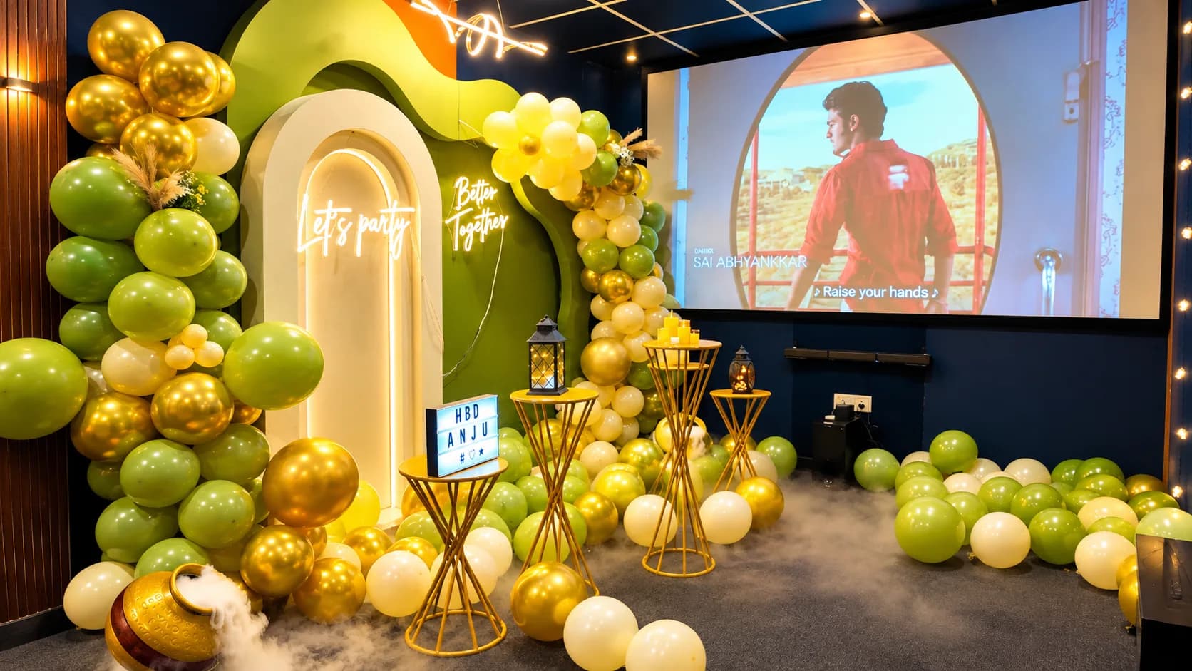 A green and gold balloon arch around a cream arch panel lit with neon Let's Party and Better Together signs, beside the projection screen playing a music video, with fog across the floor