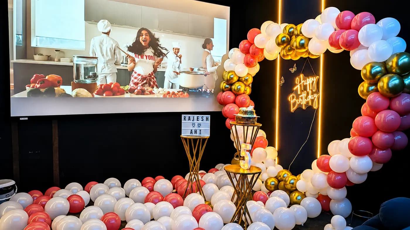 A film playing on the projection screen beside a pink, white and gold balloon ring, with a cake on a gold stand and a lit name board